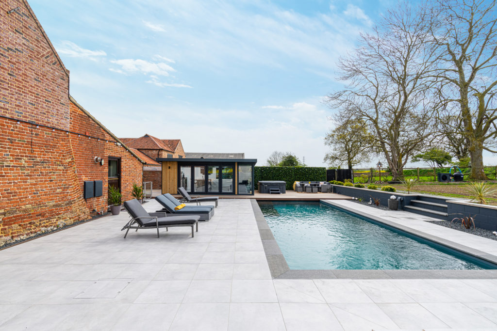 Exterior of a home bar garden building with sun loungers to the left, a hot tub and seating area to the right and a swimming pool in the foreground.