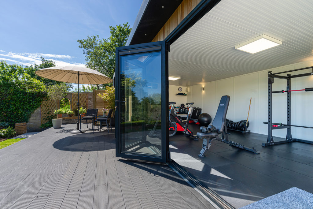 Exterior of a garden gym on a patio with a bistro set and umbrella looking into the gym with gym equipment