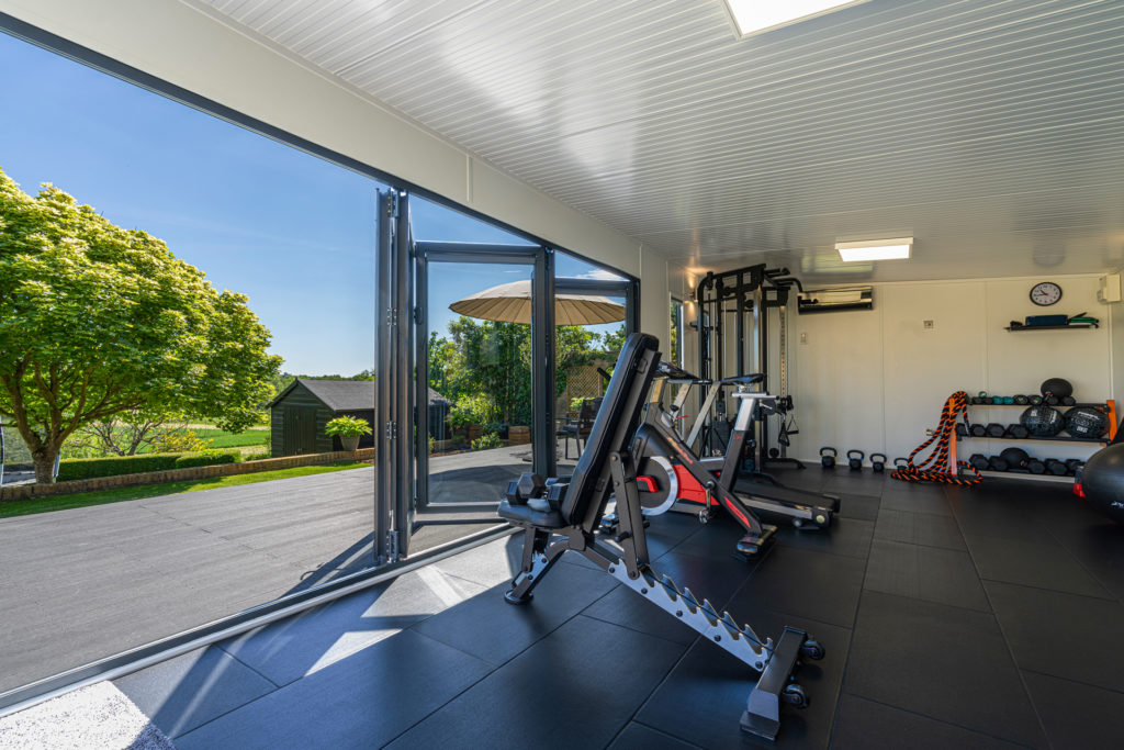 Interior of a home gym with gym equipment inside the doors are half open and looking on to a big tree and a shed